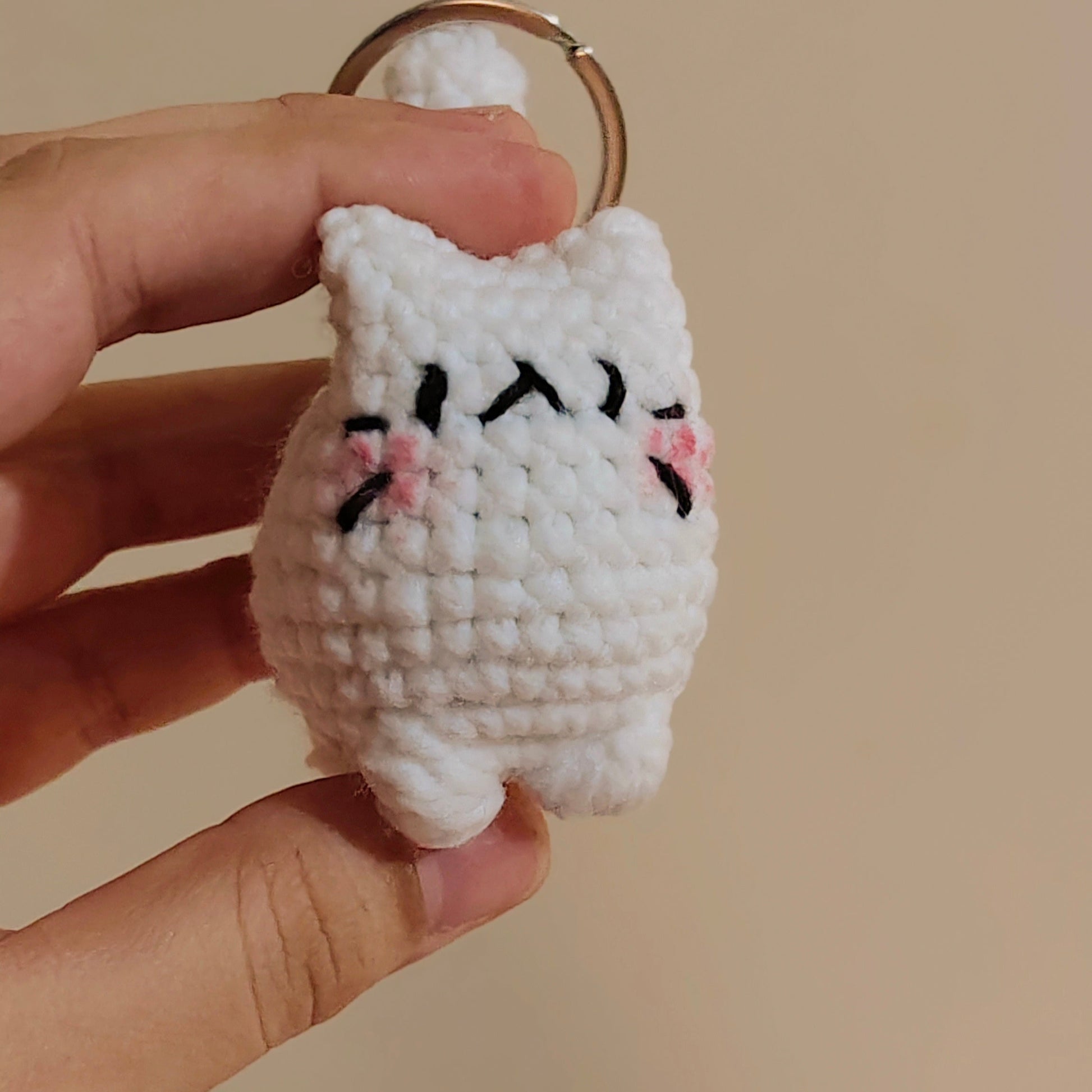 Hand holding a small white crocheted cat keychain against a beige background