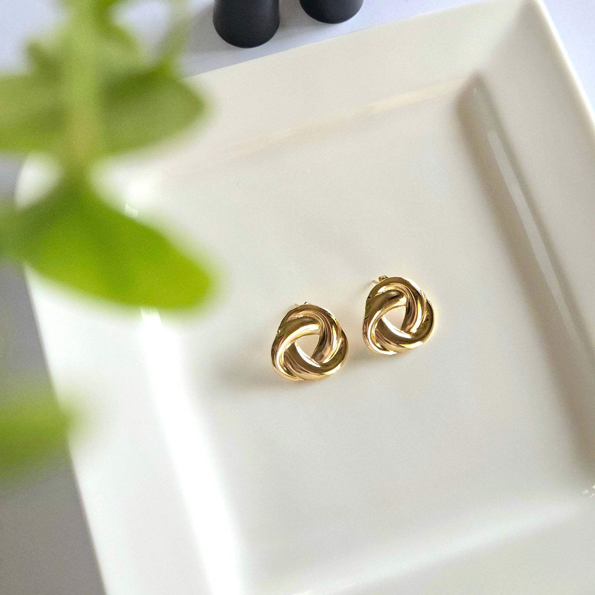 Gold knot earrings on a white dish with black lipstick and green leaves in the background