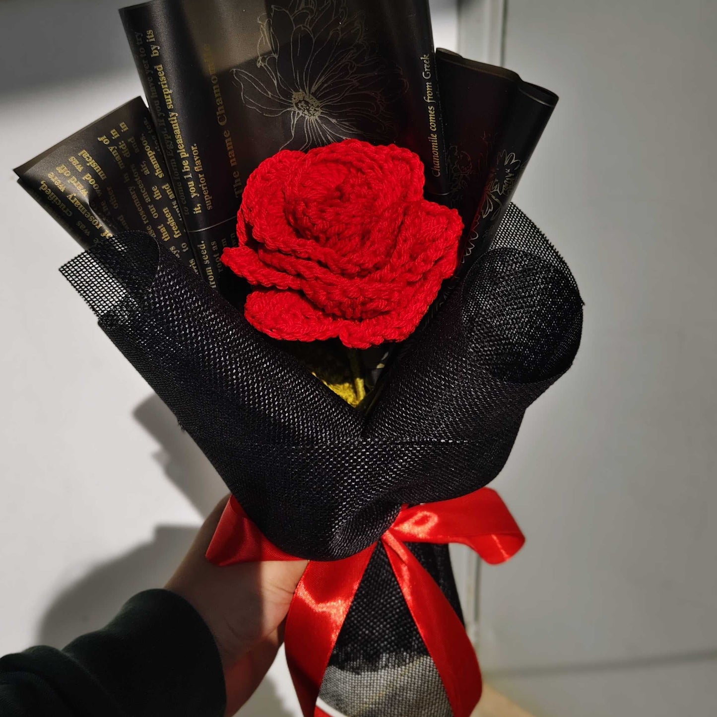 Bouquet with a red rose wrapped in black paper and red ribbon, held against a neutral background.