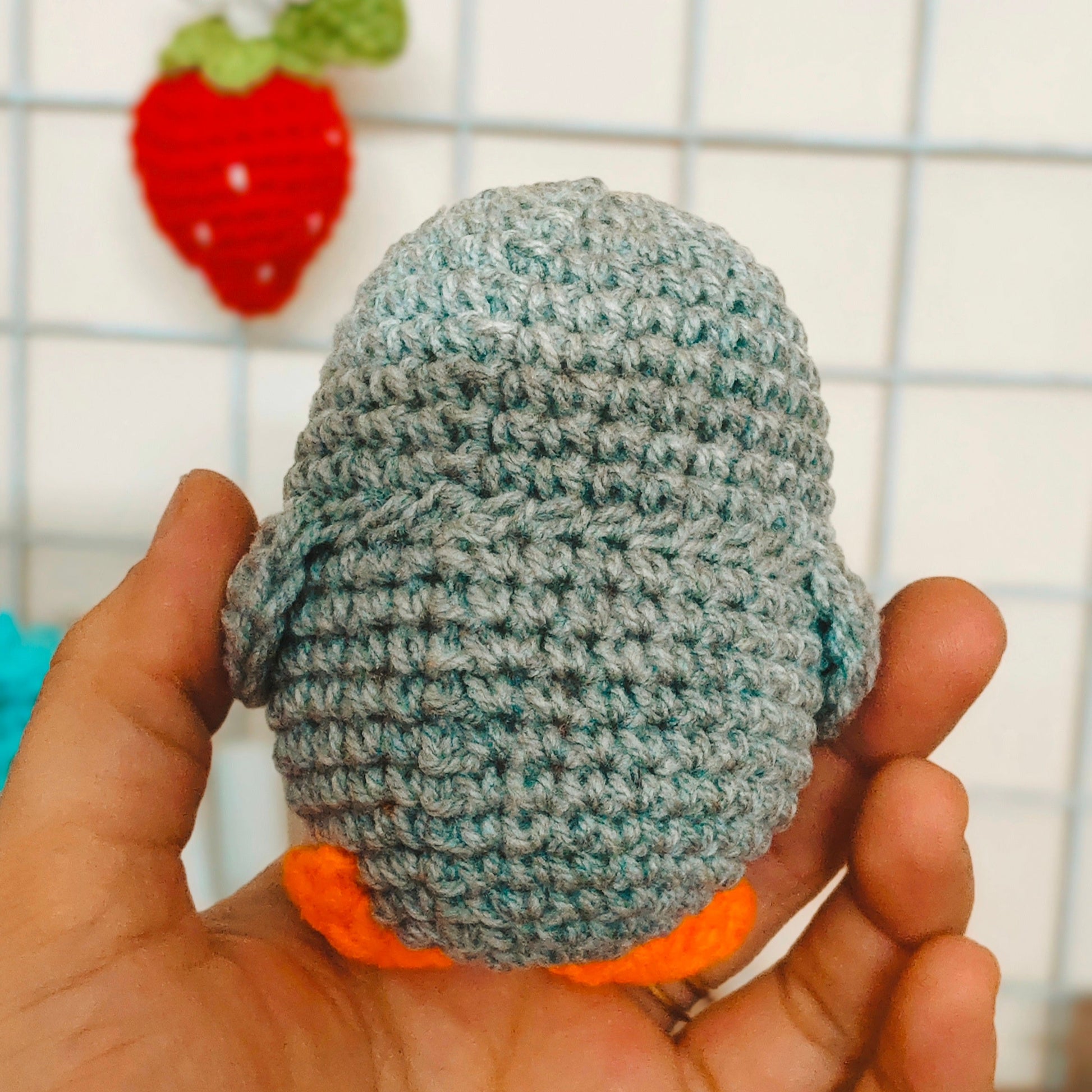 Hand holding a small crocheted gray ball with an orange bottom against a wire basket background.