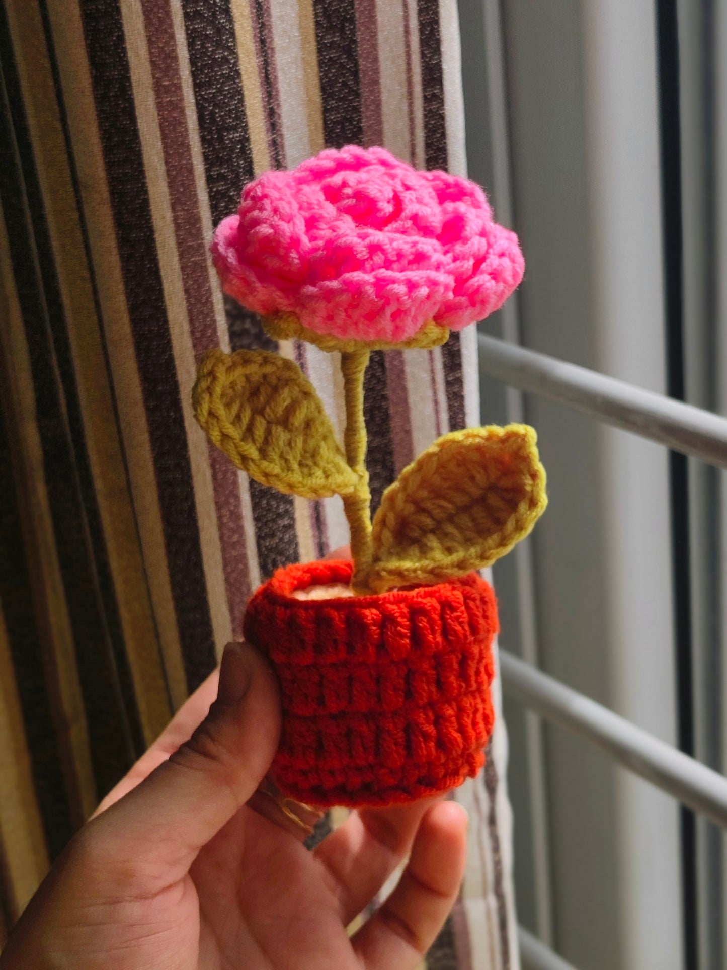 Handmade crochet pink flower in a red pot held against a striped curtain.