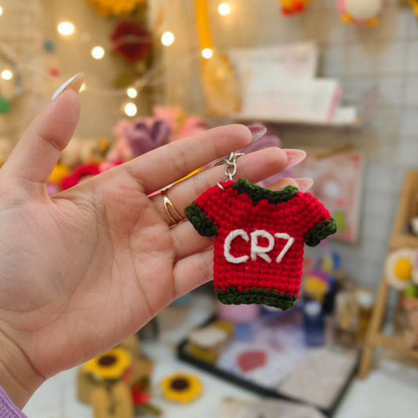 A hand holds a crocheted keychain featuring a black and white soccer ball and a red soccer jersey with 'CR7' in white, set against a softly blurred craft room background.