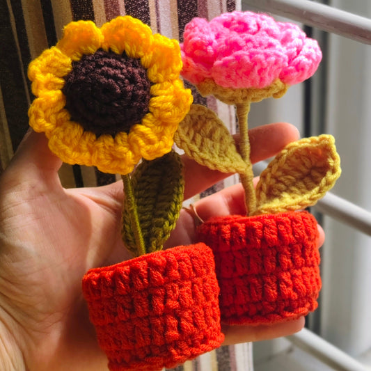 Hand holding two small crocheted flowers, one yellow and one pink, in red pots.
