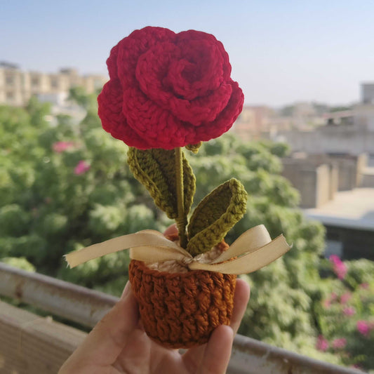 Hand holding a small crocheted red rose with green leaves against a blurred outdoor background.