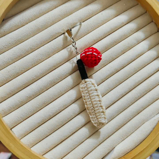 A handmade crochet keychain or bag charm displayed on a beige circular jewelry ring. The charm features a cream-colored crocheted body with a black connector and a red textured top, attached to a silver fishhook clasp.