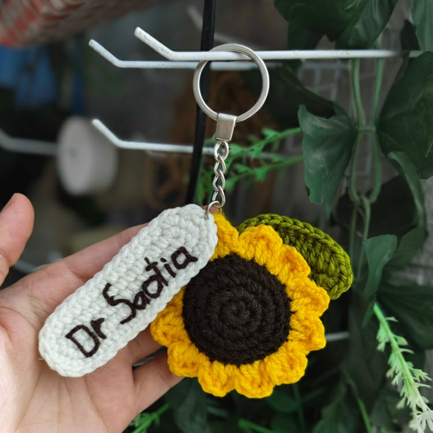Handmade sunflower keychain with 'Dr. Sadia' tag held by a hand against a blurred green background.