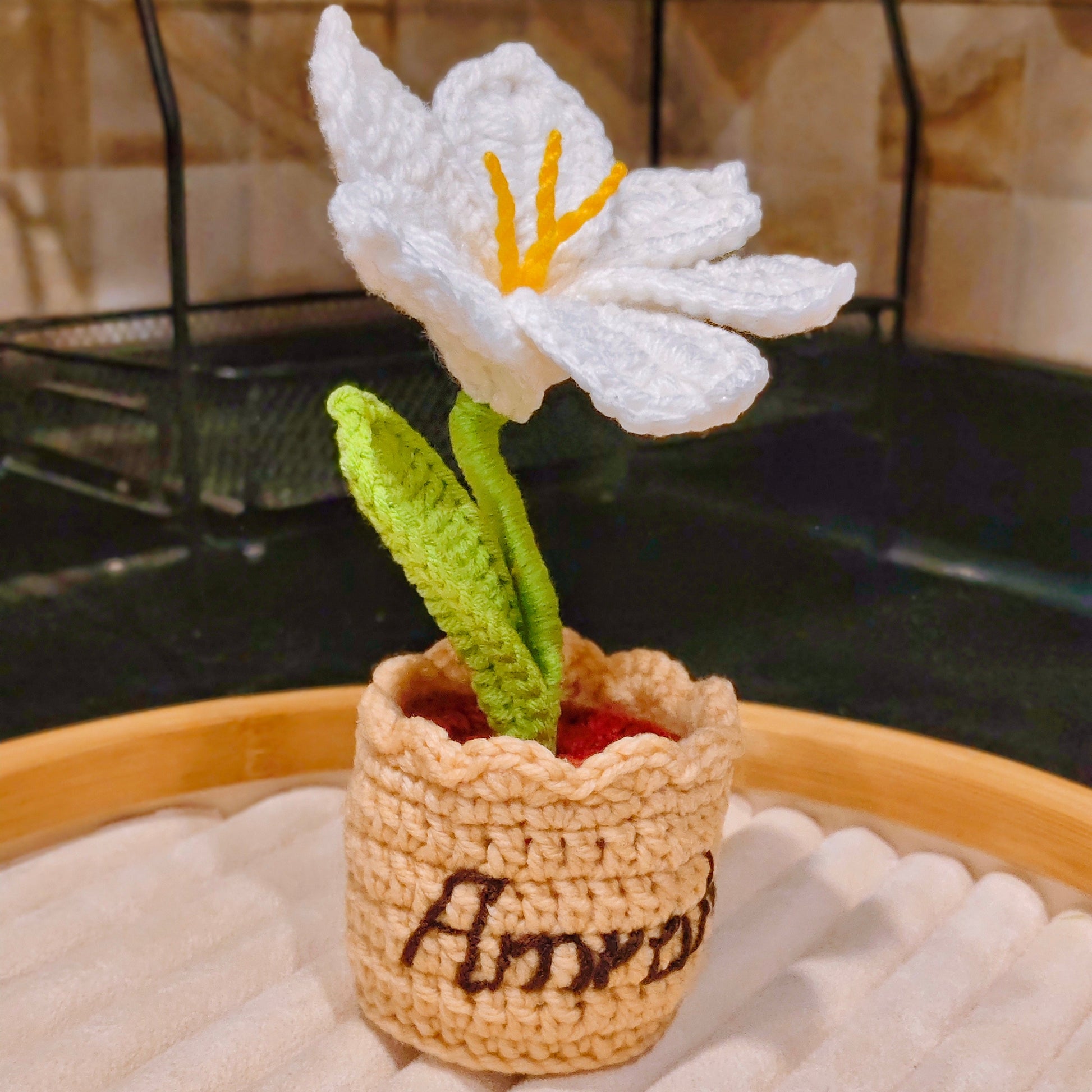 Crocheted white flower in a pot with 'Amarah' written on it, placed on a textured surface.