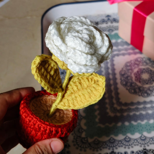 crochet white rose  flower in a pot held by a hand on a patterned surface.