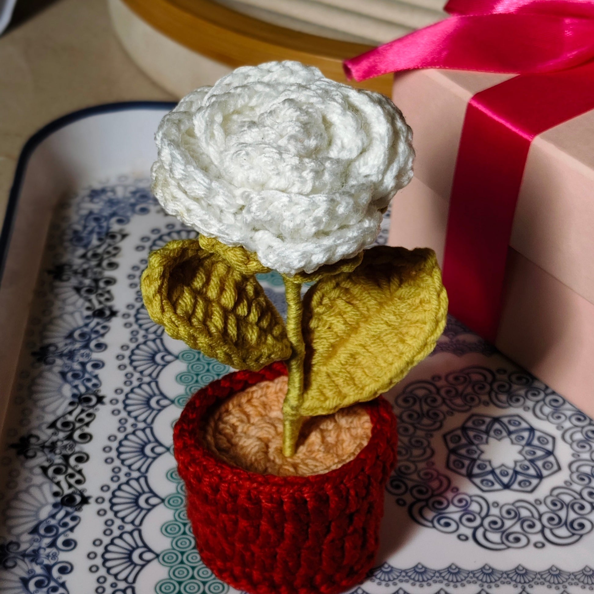 Knitted white rose in a pot on a patterned surface with a gift box in the background